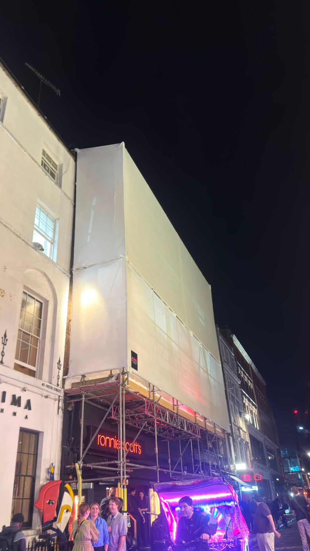 Fully encapsulated shrink wrap scaffold on Ronnie Scott's, Soho, with the club open and the public passing beneath the pedestrian gantry - Pinnacle Scaffolding